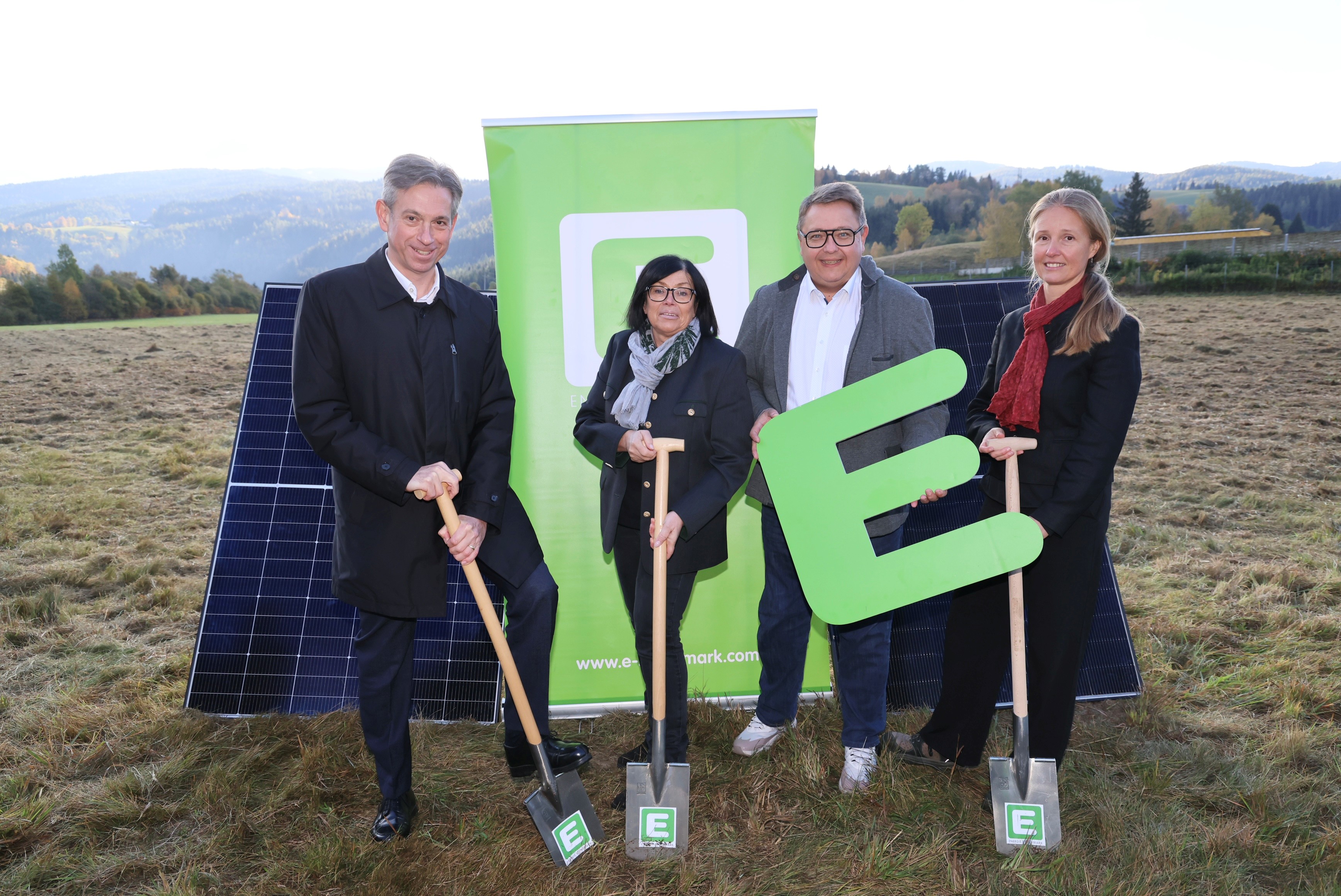 Vier Personen mit Schaufeln und einem großen grünen "E" vor Solarmodulen und einer Bergkulisse.